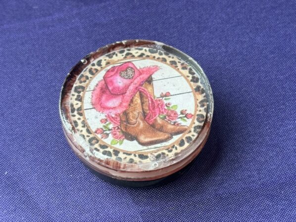 Front view of cowgirl resin phone grip with pink hat, brown boots, and leopard trim on purple fabric