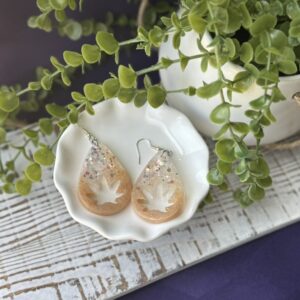 Teardrop dangle earrings with leaf shape and glitter accents, styled in a white dish beside green plant leaves.