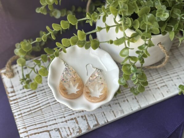 Teardrop dangle earrings with leaf shape and glitter accents, styled in a white dish beside green plant leaves.