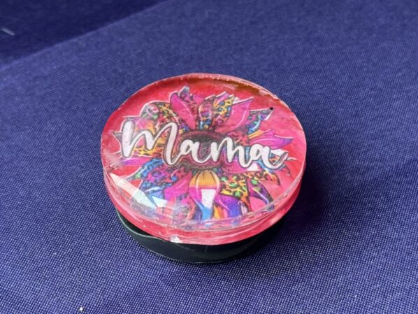 Close-up of handmade Mama phone grip featuring resin dome and multicolor glitter pattern
