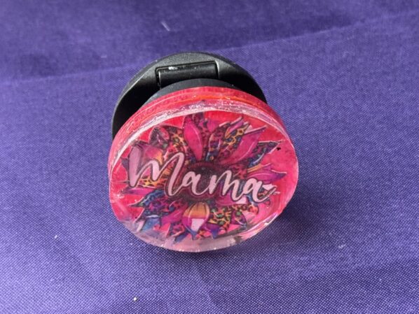 Side view of Mama resin phone grip with black collapsible base and pink glitter top, shown on purple fabric