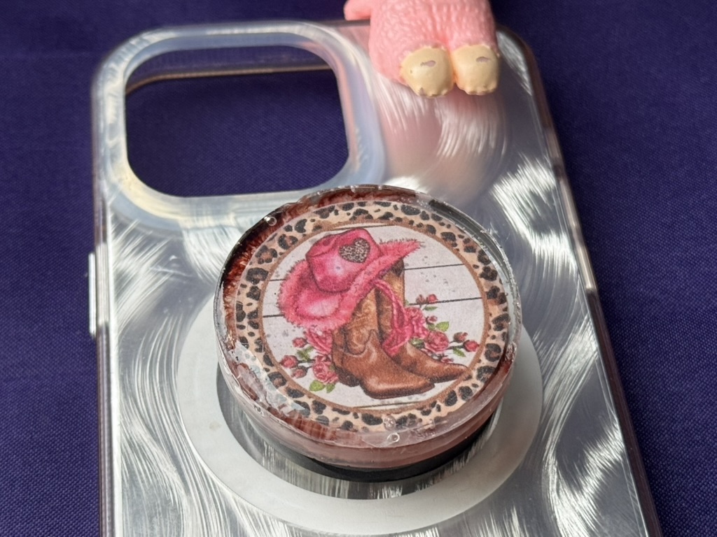 Cowgirl resin phone grip mounted on silver swirl phone case next to pink charm