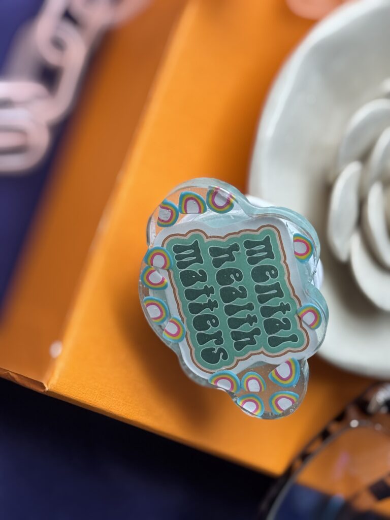 Cloud-shaped resin phone grip with “Mental Health Matters” quote, rainbow accents, and translucent blue finish displayed on an orange surface.