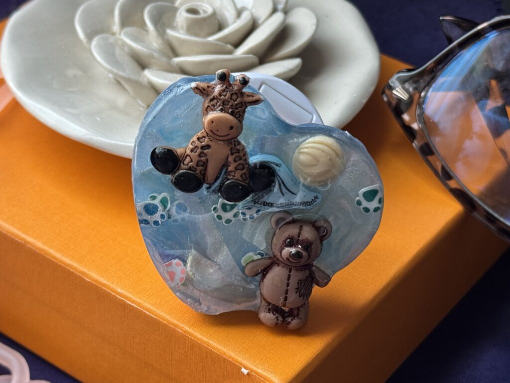 Heart-shaped blue resin phone grip with giraffe and bear details displayed on a ceramic tray with a white rose.