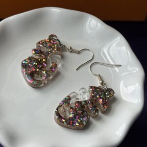 Pair of clear multicolor glitter resin earrings displayed on a soft white fur fabric. The abstract face shape earrings shine with colorful glitter under natural light, with silver hooks attached for comfortable wear. Handmade resin jewelry designed to add bold sparkle to any outfit.