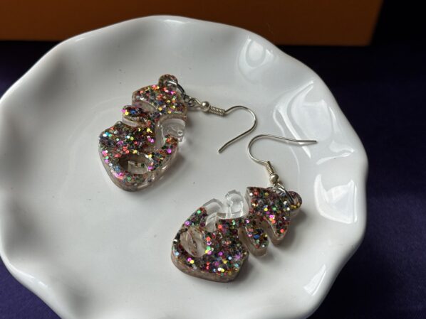 Pair of clear multicolor glitter resin earrings displayed on a soft white fur fabric. The abstract face shape earrings shine with colorful glitter under natural light, with silver hooks attached for comfortable wear. Handmade resin jewelry designed to add bold sparkle to any outfit.